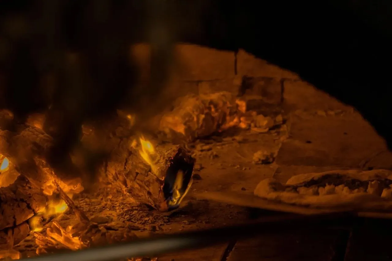 A pizza on a peel inside the wood oven with the fire burning at the back of the dome