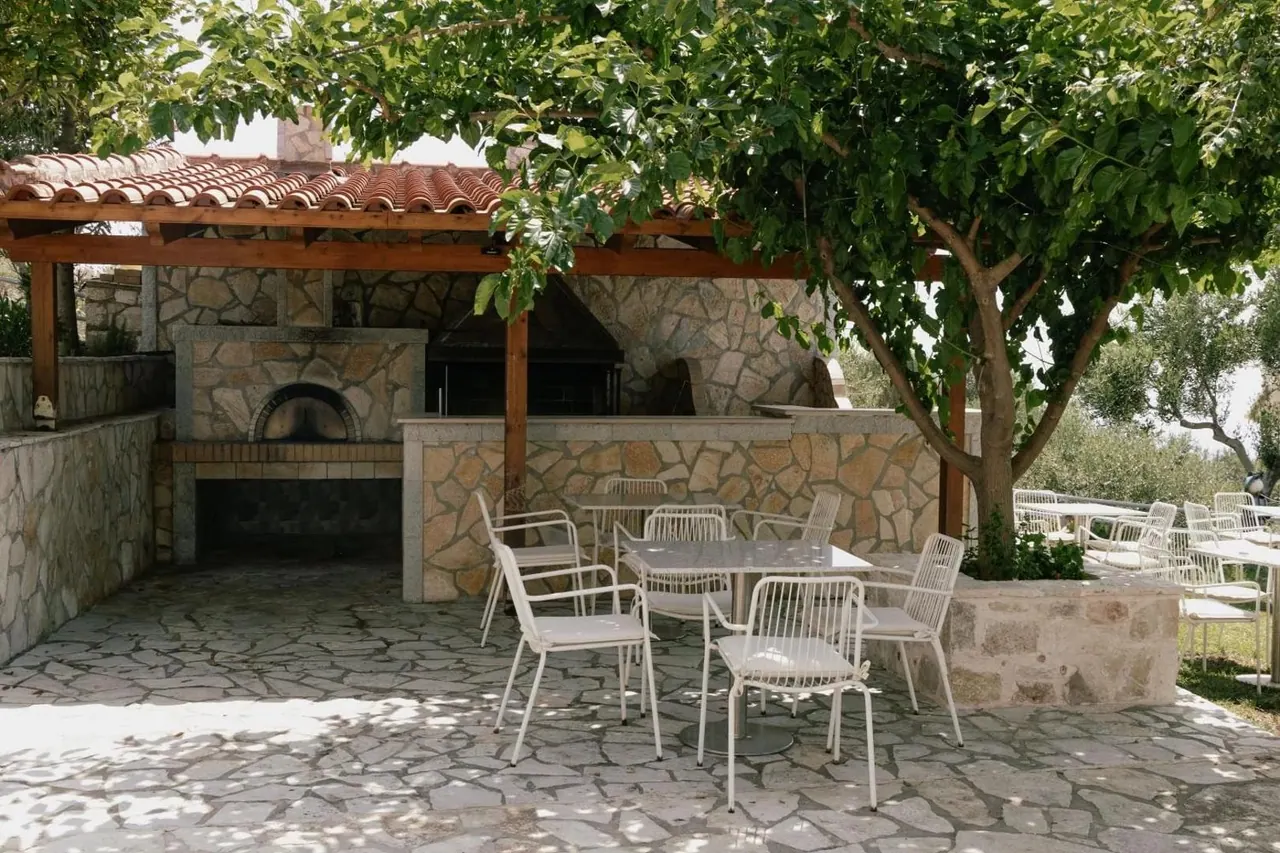 The oven and its stone counter in daylight with white chairs and tables under a fig tree