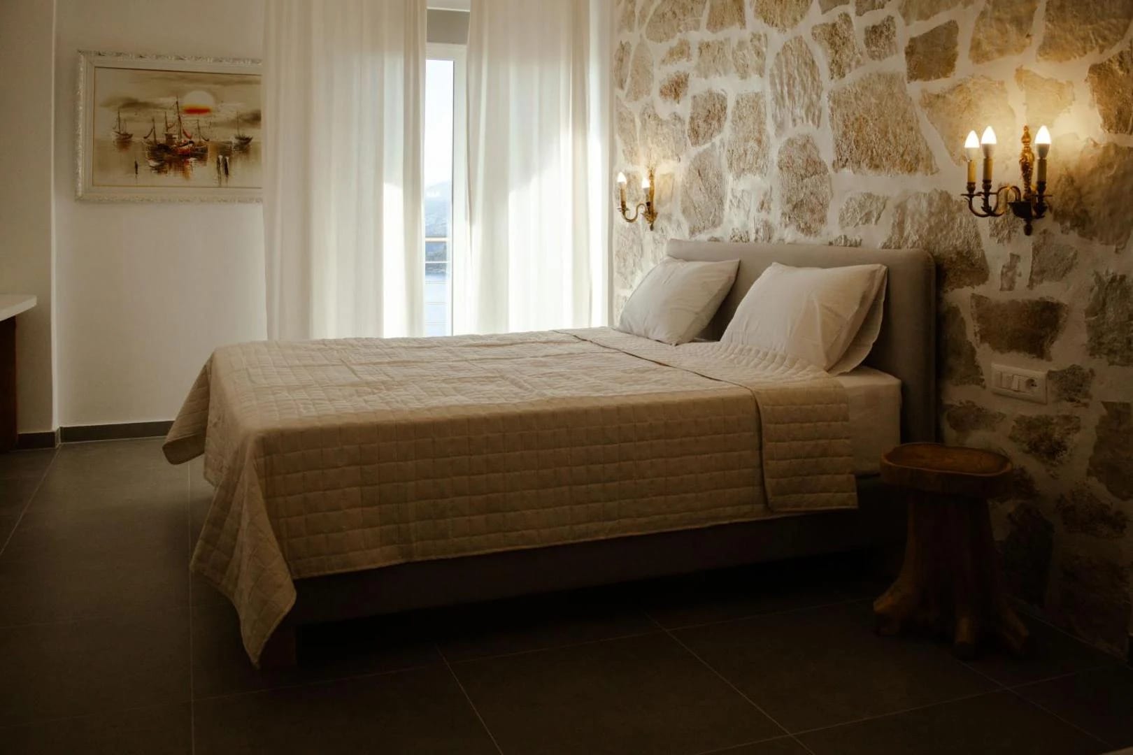 A double bed with a linen headboard against a stone wall, a window on the right looking out to the sea
